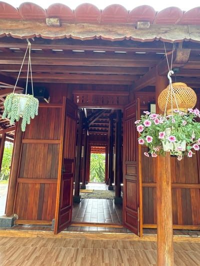 The image depicts a covered walkway in Liên Nghĩa, Vietnam, characterized by dark wood construction. The walkway extends into the distance, culminating in an opening that reveals a green landscape. Above, the wooden ceiling is interrupted by a series of cabinets with doors that are partially open. Two hanging plants adorn the space, one with pink flowers and the other with succulents. The walkway transitions to a tiled surface on the right side of the frame.