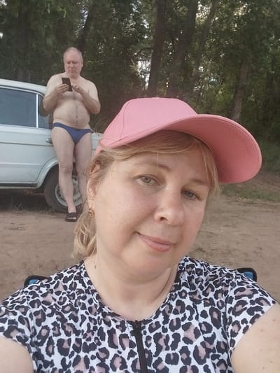 In Chkalov, Russia, a woman in a pink baseball cap and a leopard-print top is taking a selfie. Behind her, a man wearing blue briefs and holding a smartphone stands by a white car, with trees and foliage forming the background. The ground appears to be dirt.