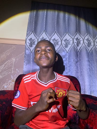 A young Nigerian boy wearing a red Manchester United soccer jersey with white trim on the collar and sleeves sits on a red sofa. The jersey features the Manchester United crest on the left chest and the Premier League logo on the left sleeve. The boy is looking upwards and to his left, with his fists clenched in front of his chest. The background consists of a patterned grey curtain and a wall with white paint splatters. A bright light casts a distinct shadow on the wall and curtain. The setting is indoors, likely in a home in Barakin Sabawa, Nigeria.