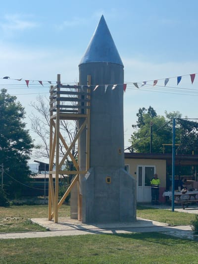 The image depicts an outdoor scene in the city of Zîrnești, Moldova, featuring a conical structure resembling a rocket, possibly a playground element or a decorative object, with a wooden scaffolding attached to its side. Small triangular flags in red, white, and blue are strung across wires above the structure, suggesting a festive or event-related atmosphere. In the background, a light-colored building with white doors and windows is visible, with a man in a bright yellow and green vest standing in front of it. Several picnic tables with checkered tablecloths are set up in front of the building, with some people seated at them. Trees and green grass form the surrounding environment, with a clear blue sky overhead.