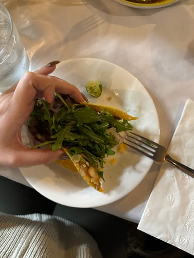 The image displays a close-up, top-down view of a dining scene within a restaurant in Guadalajara, Mexico. A fair-skinned hand, featuring long, brown-polished fingernails, grasps a partially consumed taco with a yellow corn tortilla. The taco's filling is predominantly green arugula, interspersed with white elements consistent with chopped onion or cheese, and underlying meat or seafood. The taco is positioned over a white circular plate, which also contains a squeezed lime wedge and scattered food remnants. A silver-colored, four-tined fork rests on the plate's right side. In the upper-left quadrant, a clear glass filled with water and ice is visible. A crumpled white paper napkin with an embossed leaf or feather pattern is partially visible in the lower right. All items are situated on a white tablecloth, which shows a distinct shadow of a fork in the upper-mid portion of the frame. The lower left displays a segment of a light-colored, ribbed garment worn by the individual.