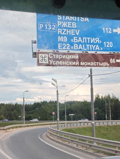 The image shows a road sign with directions. The sign is blue and has white lettering. The text on the sign is in Russian and English. The Russian text says "СТАРИЦКИЙ УСПЕНСКИЙ МОНАСТЫРЬ", which translates to "Staritsky Uspensky Monastery". The English text says "STARITSA", "Rzhev", "M9 <<BALTIYA>> 120", and "E22 <<BALTIYA>> 120". There is also a smaller brown sign with a symbol that looks like a stylized H. The road is a highway with multiple lanes and guardrails. There are trees and green grass on the side of the road. The sky is cloudy. The image is taken from inside a vehicle, as evidenced by the blurry foreground and the reflection in the upper part of the image. The location is in the city of Glinkovo, Russia.