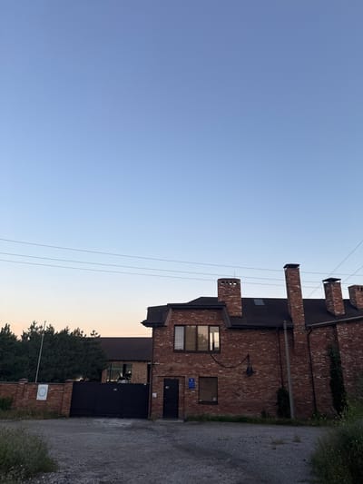 The image shows a large, two-story brick building with multiple chimneys, located in Obukhivka, Ukraine. The building has a dark roof and several windows. In front of the building is a black metal gate and a brick fence. Behind the fence, there are trees. The sky is clear and blue, with some power lines visible. The scene appears to be a private residence or a business establishment. There are no people visible in the image. The ground in front of the building is covered with gravel.