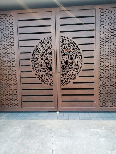 The image shows a close-up of a closed brown metal gate with intricate designs. The gate is divided into two panels, each featuring horizontal slats. The left and right sides of each panel are decorated with a repeating geometric pattern of interlocking hexagons. In the center of each panel is a large circular cutout with a complex, possibly historical or cultural, motif. The gate appears to be in a residential or commercial setting, with a grey tiled walkway in front of it and a concrete surface beyond. The lighting suggests it is daytime, with some ambient light reflecting on the gate. No people or actions are visible. The context of Lam Vien - Da Lat, Vietnam, is not visually verifiable from the image itself.