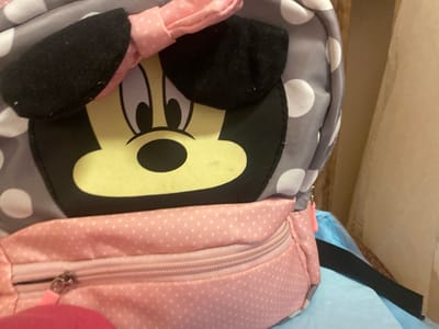 A close-up, eye-level shot focuses on a gray and white polka dot backpack with Minnie Mouse ears and a pink bow on top. The backpack features a large, yellow Minnie Mouse face with black eyes and nose. Below the face is a pink zippered pocket with a small zipper pull. The backpack is leaning against a light blue surface, and a tan curtain or wall is visible in the background to the right. There are no people or animals present in the image. The scene does not depict any identifiable landmarks or features of the city of Afanasyevo, Russia.