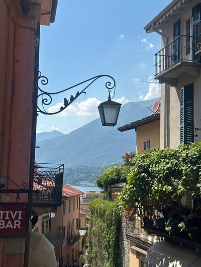 The image depicts a narrow street scene in San Giovanni, Italy, under a clear blue sky with scattered white clouds. The perspective is tilted, giving a sense of looking up and through the street. Buildings with traditional architecture line the street, featuring shuttered windows, balconies with ornate metal railings, and terracotta roof tiles. Lush green foliage spills over from balconies and along the side of one building, adding a touch of natural beauty to the urban environment.
In the foreground, an antique-style lamppost with intricate scrollwork is a prominent object. Beyond the buildings, a majestic mountain range dominates the background, its slopes covered in dense greenery. The overall atmosphere is serene and picturesque, suggesting a pleasant daytime visit.
One building displays a sign that reads "TIVI BAR," indicating a commercial establishment. The time of day appears to be midday, given the bright sunlight casting shadows and illuminating the scene. The weather is fair and sunny, conducive to outdoor activities. The scene captures the charm and beauty of a European town, with a blend of historical architecture and natural landscapes. No people are clearly visible in the image, but the presence of the bar sign suggests it might be a place where people gather. The overall impression is one of peaceful exploration and appreciation of the local scenery and culture.