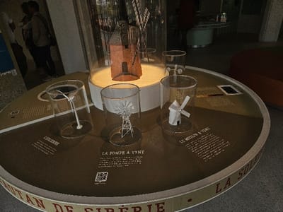 This image depicts an indoor exhibit within the Maison de la Haute-Garonne - Canal du Midi in Avignonet-Lauragais, France. The central element is a large, circular, dark brown display table with a lighter outer rim that includes partial French text. At its core, a detailed model of a red-brick tower windmill with a dark roof stands under a tall, cylindrical clear enclosure, illuminated from below. Surrounding this central model are four smaller cylindrical clear cases, each containing a different model related to wind power: a white vertical-axis wind turbine labeled "L'ÉOLIENNE," a multi-bladed wind pump on a skeletal tower labeled "LA POMPE À VENT," a white traditional horizontal-axis windmill labeled "LE MOULIN À VENT," and a small cup anemometer. French explanatory texts and QR codes are printed directly onto the display table's surface adjacent to each exhibit. A small, dark rectangular object, possibly a tablet, lies on the table to the far right. In the blurred background on the left, two individuals are partially visible, one in a light-colored top, the other in a darker garment, standing near a vertical display panel. A third indistinct figure is partially visible further back on the right. The floor is dark, and the walls are light-toned.