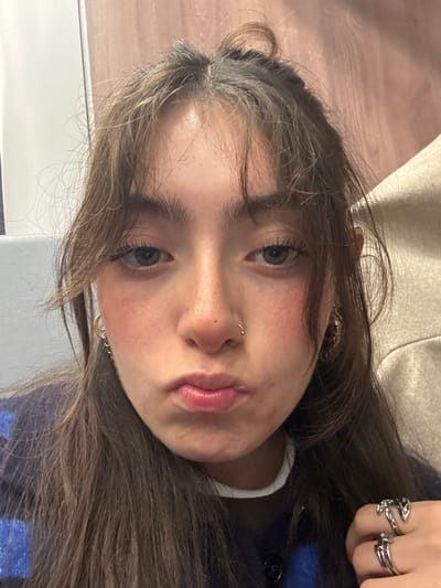 A youthful person, possibly a teenager, with light brown hair and fair skin is viewed in a close-up, slightly angled shot. They have green eyes and a nose piercing. The person is wearing multiple silver rings on one hand. They appear to be pouting with glossy lips. Background elements include a beige fabric and a blue and white surface.