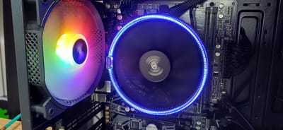 This is a close-up, eye-level, indoor shot of the interior of a computer case. On the left, a large computer fan with colorful RGB lighting is visible. To its right, another fan, likely a CPU cooler, is mounted on a motherboard. This fan has a blue LED ring around its edge and a white logo, possibly "PCCooler," visible on its side. The motherboard itself is mostly black with various components and black cables connected to it. The computer case is black and appears to be made of metal and plastic. The background is dark and out of focus. There are no people or animals present in the image. The scene is well-lit, likely by the RGB lighting of the fans. There is no information in the image to suggest a location in Zuevka, Russia.