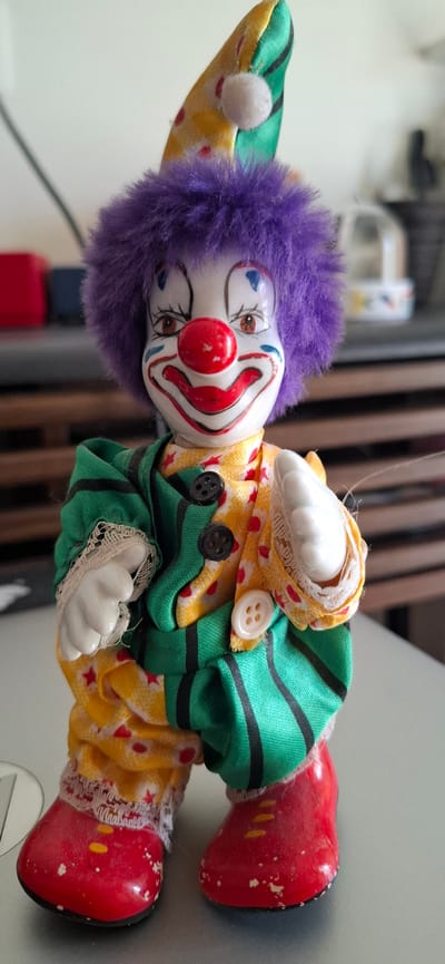The image shows a close-up, sideways view of a porcelain clown doll lying on a flat surface. The doll is dressed in a colorful costume. It has a white face with traditional clown makeup, including a red nose, painted red lips, and blue accents around the eyes and eyebrows. The doll has bright purple, fuzzy hair and wears a pointed hat with green and yellow stripes. Its body is covered by a green shirt with black stripes and yellow pants decorated with red stars. White ceramic hands and red, pointed shoes complete the outfit. Two black buttons and two white buttons are visible on the doll's clothing.
The doll appears to be a marionette or string puppet, as a thin white string is visible attached to its head, extending upwards and out of frame. The background is blurred, but suggests an indoor setting with vertical wooden elements, possibly a rack or blinds, and a light-colored wall. There is no discernible text in the image. The lighting suggests it is daytime. The image is static, showing the doll at rest.