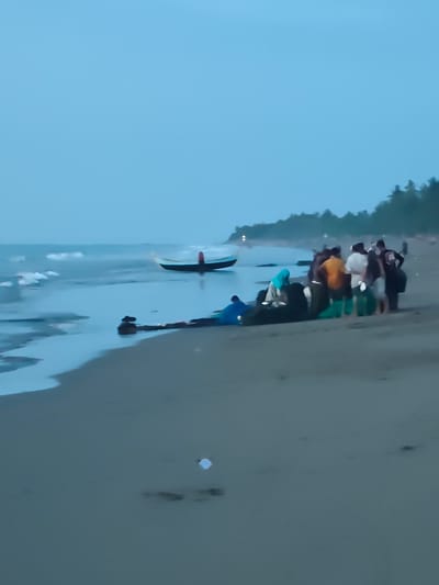 A group of people are gathered on a sandy beach in Curehtunong, Indonesia, near the shoreline. The sky is overcast and the sea is calm with small waves. A traditional fishing boat is beached on the sand, and several individuals are either standing around it or interacting with it. Some of the people are wearing light-colored clothing, while others are in darker attire. The environment suggests a coastal setting with a line of trees visible in the distance. The overall scene appears to be a moment of activity related to the fishing boat on the beach.