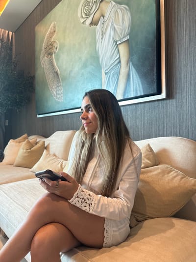 An interior scene in Monterrey, Mexico, depicts a young woman seated on a light-colored sectional sofa. The woman, with long light brown hair, is dressed in a white long-sleeved top featuring white crochet lace cuffs and white shorts with a matching lace trim. She is positioned with her legs crossed, holding a black smartphone in both hands, and wears a ring on her left ring finger. Her gaze is directed towards the right side of the frame with a slight smile. Behind her, a large rectangular painting hangs on a dark gray, vertically striped wallpapered wall. The artwork portrays an owl with open wings on the left and a stylized female figure with a large white rose covering her head on the right, against a muted greenish-blue background. To the far left, a dark green potted plant is partially visible. The sofa is adorned with multiple light-colored pillows, some exhibiting a subtle wavy texture.