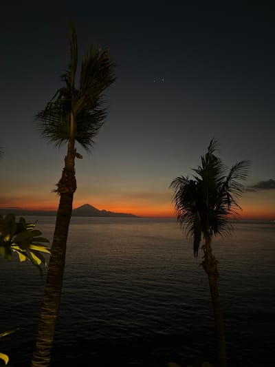 The image shows a serene dusk scene with two silhouetted palm trees in the foreground, framing a body of water. The sky transitions from a deep blue at the top to a vibrant orange and yellow gradient near the horizon, indicating either sunrise or sunset. In the distance, a conical mountain, likely Mount Rinjani, is visible, rising above the horizon. The water's surface is calm, reflecting the sky's colors. The scene is captured from a high vantage point, possibly a balcony or cliffside, overlooking the ocean. Two faint celestial bodies are visible in the upper portion of the dark sky. The overall atmosphere is tranquil and picturesque. This view is characteristic of the coastline in Senggigi, Indonesia, known for its beautiful sunsets and views of Mount Rinjani.