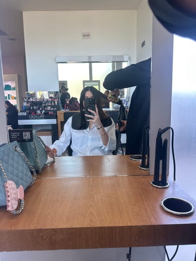 A woman is reflected in a salon mirror, taking a selfie with her phone. A hairstylist is working on her hair. On the counter, next to the mirror, is a blue quilted purse with a price tag and a pink hair clip on top. Behind the woman, shelves filled with beauty products can be seen, and another person is visible in the background.
