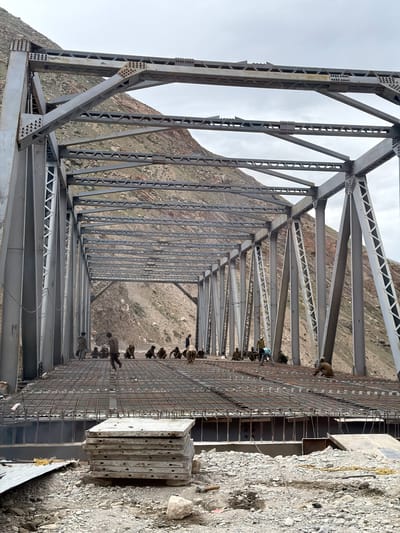 The image shows the construction of a steel truss bridge over a rocky, mountainous terrain. The bridge structure is largely complete, with workers on the deck preparing for concrete pouring. The deck is covered with a grid of rebar, and several workers are visible sitting or standing on it. In the foreground, a stack of precast concrete slabs is piled on the ground. The background features steep, barren mountain slopes under a cloudy sky. The scene appears to be in a remote, possibly underdeveloped area, indicated by the rough terrain and the method of construction. There are approximately 15-20 individuals visible, appearing to be construction workers, engaged in various tasks related to the bridge construction. The overall environment is dusty and rocky, consistent with a construction site in a mountainous region.
