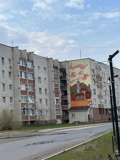 A high-angle shot shows a tall, grey, brick building with a large mural on the side, located in Pokrov, Russia. The mural features a stylized cityscape with red and brown buildings, a tall steeple, and abstract figures in red and yellow. The building has numerous windows, many with satellite dishes attached. A decorative metal street lamp is visible in the foreground. The road runs alongside the building, and a patch of green grass and some bushes are visible. The sky is overcast with light blue and grey clouds. Puddles are visible on the road surface.