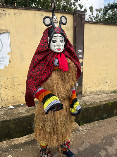 In Alagba, Nigeria, a costumed figure stands outdoors against a textured yellow wall. The figure wears a large, straw-like costume with brightly colored knitted sleeves. A red and black patterned fabric drapes over the shoulders, and a red scarf is visible around the neck. The headpiece is elaborately decorated with two figures, one larger with circular elements, and a smaller, human-like face. The lower part of the costume has decorative elements and dark blue pants. A wooden stick is held by the figure. The environment includes a concrete drainage ditch and a paved surface. A partial sign is visible on the wall.