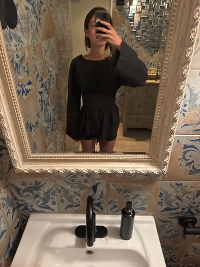 An individual is depicted taking a mirror selfie in a well-appointed restroom. The person, appearing female, wears a dark-colored, long-sleeved romper with a cinched waist and wide sleeves, obscuring her face with a silver-colored smartphone held in her right hand. She is reflected in a large, rectangular mirror featuring an ornate, off-white frame.
The restroom walls are covered with aged-looking ceramic tiles displaying faded blue and muted green floral and botanical patterns on a beige background. In the foreground, below the mirror, is a white ceramic sink with a modern, matte black gooseneck faucet. A matching matte black cylindrical pump bottle rests on the sink's surface to the right of the faucet. A matte black wall-mounted towel bar is also visible to the right.
The reflection in the mirror reveals a background behind the individual, including a light-colored wooden chest of drawers or vanity. Above this furniture, a textured wall art piece composed of numerous small, reflective, square elements is installed. Small, indistinct objects are visible on the surface of the chest of drawers.
The precise location within the city of Miami Beach, United States, cannot be determined from the visual information provided. The environment suggests a stylish commercial establishment or a private residence.