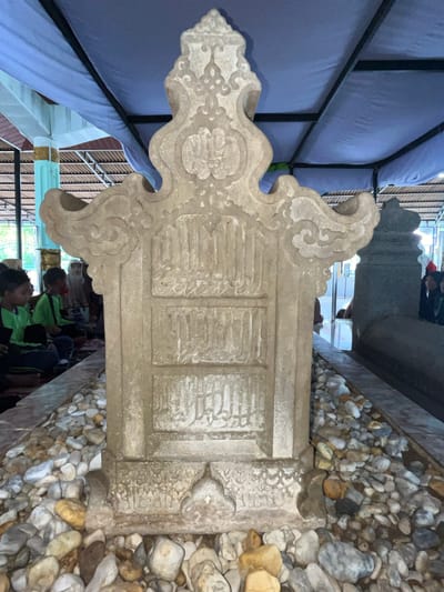 In Beuringen, Indonesia, a weathered stone tomb stands prominently in the foreground, surrounded by small, assorted pebbles. The tomb features ornate carvings and inscribed text, suggesting it is of historical or religious significance. Behind the main tomb, another, larger stone tomb is partially visible. Several people, including children and adults, are seated on the ground to the left of the central tomb, appearing to be in a solemn or contemplative state, possibly engaging in prayer or remembrance. They are partially covered by a blue tarp that extends overhead, providing shade. The environment appears to be an outdoor or semi-outdoor sacred site, possibly a cemetery or shrine, with architectural elements of a building visible in the background, including pillars and roofing structures. The lighting suggests it is daytime, with natural light filtering through the covered area.