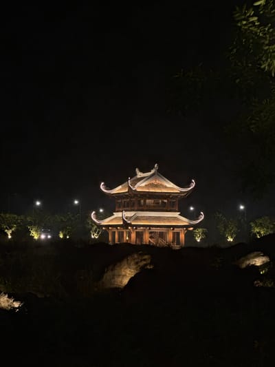 The image shows a pagoda at night in Tây Hoa Lư Ward, Vietnam. The pagoda is illuminated with warm, golden lights, highlighting its traditional architectural features, including tiered roofs with upturned eaves and intricate wooden detailing. It is situated on a slightly elevated, dark landscape with rocks and sparse vegetation visible in the foreground. In the background, several streetlights and scattered trees are visible, also illuminated by artificial lights, creating a softly lit, atmospheric scene against the dark night sky. A car with its headlights on is also visible in the distance.