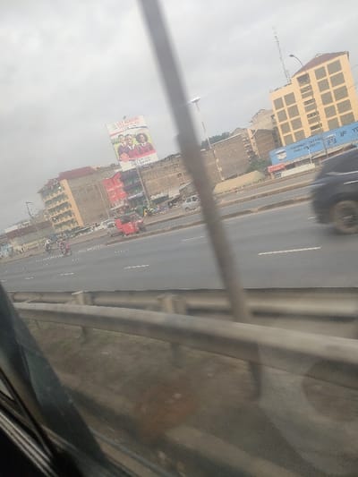 The image depicts a multi-lane urban road in Githurai, Kenya, viewed from a slightly elevated position, possibly from a moving vehicle. A metal guardrail is blurred in the foreground, with a diagonal dark element partially obscuring the bottom left. A blurry vertical pole is prominent in the mid-right.
The asphalt road features white dashed lane markings. Traffic includes a heavily blurred dark vehicle in motion on the right, a stationary or slow-moving red tuk-tuk, a light-colored sedan, and multiple motorcycles, some with passengers, on the left side of the road.
In the background, a series of multi-story buildings line the thoroughfare under an overcast sky. A tall, light-brown building with square window cutouts and a dark-tiled roof is visible on the far right. A prominent billboard advertises "LILY ACADEMY JUNIOR SCHOOL," displaying images of two smiling students in red blazers. Below this, various commercial structures are visible, including one with a red roof and another featuring a blue sign that reads "MOMBAST TEXTURE PAINT ON EARTH."