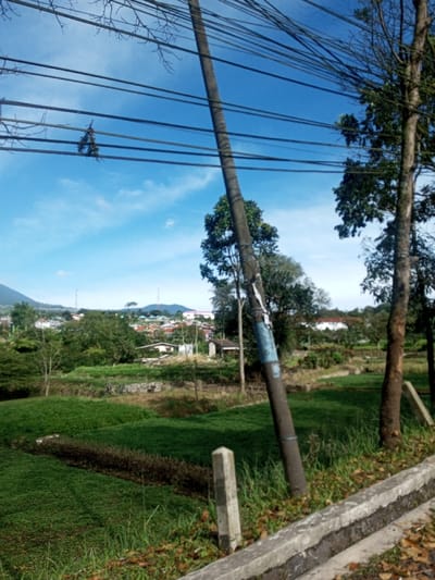 The image depicts an outdoor scene in Phonengam, Laos, under a clear blue sky with scattered white clouds. In the immediate foreground, a concrete curb or low barrier runs horizontally along the bottom right. Adjacent to it on the left, a light-colored rectangular concrete marker post is partially visible amidst green grass and scattered dry leaves. Dominating the central foreground is a dark utility pole, slightly leaning to the right, with a blue and white label or wrapping affixed to its upper section. Numerous black utility wires extend horizontally across the upper half of the frame, some intertwined with bare tree branches, and a small cluster of green foliage is visibly snagged on one of the lower wires. To the right of the utility pole, two trees with dark bark are present. The middle ground consists of expansive green fields, possibly cultivated, with a darker, narrow strip of vegetation running through them. Beyond the fields, a semi-urban settlement with numerous houses and buildings is visible, interspersed with trees. In the background, a large, green mountain range rises on the left, while several communication towers are discernible on distant elevated terrain within the settlement. No individuals or animals are present in the scene.