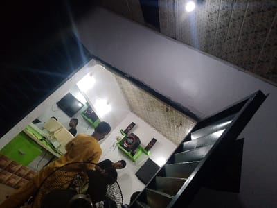 A tilted view shows an indoor space with several people. A man in a mustard-colored shirt stands near a large fan. Several people are visible behind a green counter space. A TV hangs on the wall. Speakers are positioned throughout the room. A dark staircase leads upwards along a white wall. The ceiling is visible in the upper part of the frame.