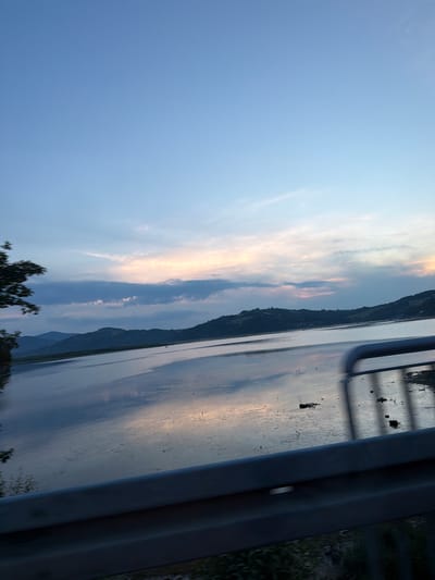 The image depicts a tranquil natural scene, not located in Sienna, Poland, as the prompt suggests. The photograph captures a wide expanse of water, likely a lake or a calm sea, reflecting the soft hues of a twilight sky. Rolling hills covered in lush greenery form a distant, serene backdrop, their contours softened by the fading light. The sky itself is a canvas of pale blues transitioning into warm shades of orange and pink near the horizon, where scattered clouds add texture and depth. In the foreground, a metallic railing suggests the perspective is from a bridge or an elevated vantage point, offering a glimpse of the natural landscape. There are no people or identifiable structures that would suggest an urban environment. The overall atmosphere is one of peacefulness and natural beauty, indicative of a quiet, undeveloped area.