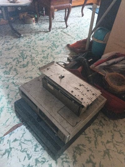 The image displays an indoor area within a building in the city of Dogsenang Gorphoog, Bhutan. The floor is covered with a light-colored surface adorned with a repeating pattern of small, teal, leaf-like designs. In the foreground, a stack of three items is visible: a dark-colored rectangular electronic device at the bottom, topped by a lighter-colored, possibly silver, rectangular electronic device featuring an access panel on its front-right. Resting on the latter is a detached silver-colored metal plate with multiple small circular perforations and slots, centered by a raised metallic fixture. To the right of these stacked electronics, a collection of objects is aggregated, including a light blue plastic bucket or bin, a light-colored pole, items of red fabric or plastic, and a brown woven object. In the background, partially visible, are two pieces of wooden furniture with slender, turned legs; one appears dark-colored on the left, and another lighter-colored piece is positioned to the right. No individuals are present in the scene, and no specific actions are depicted.