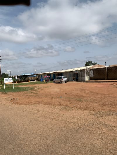 This is a ground-level, wide-angle, full shot of a building complex and its surroundings on a cloudy day in Nabatum, Nigeria. The scene depicts a commercial or administrative area with a long, low building with a white roof that appears to be a collection of shops or offices. The building has metal bars or grates over some of its windows and entrances, with blue doors and windows visible. A silver minivan is parked to the right of the building. In front of the building, there is a red umbrella, a motorcycle, and some blue gas cylinders. A brick wall forms the boundary on the right side of the complex.
To the left of the main building, there is another structure with a blue roof supported by metal poles, and some greenery underneath, possibly a greenhouse or a shaded waiting area. A white sign with text and a logo is visible in the foreground on the left, which appears to be for "JOS WATER SERVICES CORPORATION". The ground in front of the buildings is a mix of red dirt and gravel. Power lines are strung across the upper part of the image. The overall environment suggests a functional, somewhat utilitarian urban or semi-urban setting. There are no people clearly visible in the image.