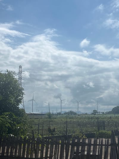 The image depicts a rural landscape in Jeneponto, Indonesia, characterized by a vast expanse of sky with scattered white clouds above a field of wind turbines. A prominent transmission tower stands to the left, its metallic structure silhouetted against the sky. In the foreground, a rustic wooden fence runs horizontally across the frame, separating the viewer from a lush green area with trees and vegetation. The background reveals a line of wind turbines, their blades visible against the horizon, suggesting a renewable energy installation. The overall scene is serene and natural, with a focus on the interplay between man-made structures and the environment.