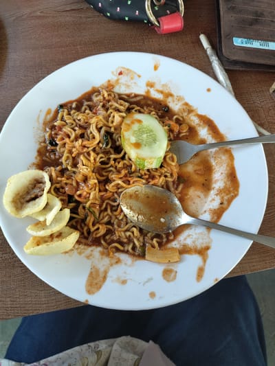 A partially eaten plate of Indonesian mie goreng (fried noodles) sits on a wooden table. The mie goreng is topped with a slice of cucumber and accompanied by some kerupuk (crackers). A fork and a spoon rest on the plate amongst spilled sauce. A wallet and a small purse with a red strap are visible in the upper right corner of the frame. The edge of dark blue trousers is visible in the lower center of the frame. The location is likely Matang, Indonesia, based on the meal.