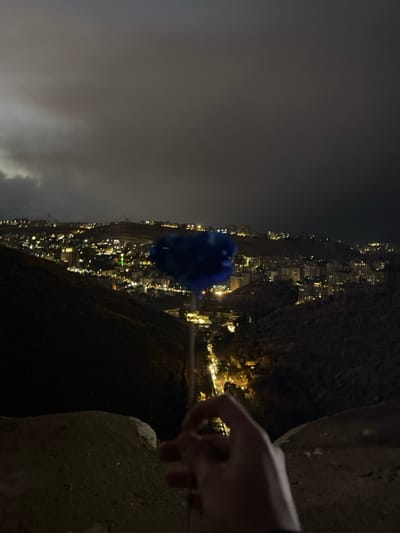 An elevated, nighttime view captures a section of Dummar Municipality, Syria, under a cloudy sky. In the foreground, a human hand is visible on the lower right, holding a slender stick-like object topped with a spherical, fuzzy blue item, possibly a piece of cotton candy or a decorative puff. The immediate ground appears uneven and unpaved. A deep, dark valley with a winding road illuminated by vehicle headlights separates the foreground from a densely built-up urban area. The city, situated on hills, is characterized by numerous illuminated buildings, with a prominent green light visible among them. The distant landscape consists of dark, undulating terrain under a heavy, obscured sky.