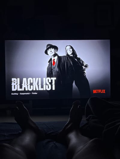 The image shows a television screen displaying the Netflix interface for the show "The Blacklist." The screen is a wide shot, with the show's title and two main characters, a man in a fedora and a woman, prominently featured. Below the title, the words "Exciting," "Suspenseful," and "Thriller" are displayed, along with the Netflix logo. The scene is dimly lit, suggesting a nighttime viewing experience. In the foreground, a person's bare feet are visible, resting on a dark blue surface, possibly a bed or couch, implying a relaxed setting. The overall impression is of someone watching television at home. The image is not specific to Mollet Del Vallès, Spain, as no identifying landmarks or contextual details of that location are present.