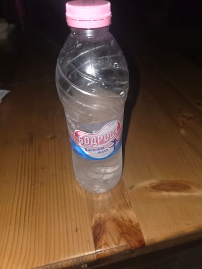 A clear plastic bottle with a pink cap rests on a wooden surface. The label on the bottle reads "Бодрост Трапезна вода" in Cyrillic, indicating it contains mineral water. Condensation droplets are visible on the bottle's exterior. The wooden surface appears to be constructed of planks. The location, Pripek, Bulgaria, is based on the product’s origin.