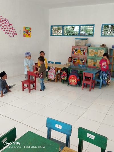 The image depicts an indoor classroom or play area in Jagapura Kulon, Indonesia, captured on October 14, 2025, at 07:34. The room features white tiled flooring and plain white walls. Two horizontally oriented windows with blue frames are visible on the far wall, providing a view of green foliage outside. Decorative red floral decals are applied to the left wall, alongside a colorful educational chart.
Seven young children, presumably preschoolers or early primary school students, are present.
* On the far left, a male child wearing a black peci and grey long-sleeved shirt sits on the floor with his legs extended, looking towards the center.
* In the left-center, a male child wearing a light blue patterned shirt and a blue kopiah stands, holding the back of a red child-sized chair with his left hand, looking down at another child.
* Adjacent to him, another male child in a brown patterned shirt and blue jeans stands, facing left, with his left hand resting on the seat of the red chair.
* Behind these two, a female child in a yellow long-sleeved shirt stands partially obscured, observing the scene.
* Towards the right, a male child in a light-colored shirt is seated at a wooden child-sized table, with a clear bottle on the table in front of him.
* Further right, a female child in a pink short-sleeved shirt stands behind a small red table, her right hand near her face.
* Another female child is partially visible near a table in the mid-left area.
The room is furnished with several child-sized wooden tables and chairs, painted in varying colors such as light blue, green, and red. Two chairs in the foreground, painted green and blue, have labels "28" and "4" taped to their backs. Along the far wall, two sets of multi-compartment cabinets, decorated with cartoon characters (Mickey Mouse on the left set), are visible. A clear plastic storage container rests on top of the left cabinet. Multiple child-sized backpacks with various designs, including Spider-Man and Iron Man, are positioned near the tables and chairs. The scene suggests an active environment with children interacting within a designated learning or play space.