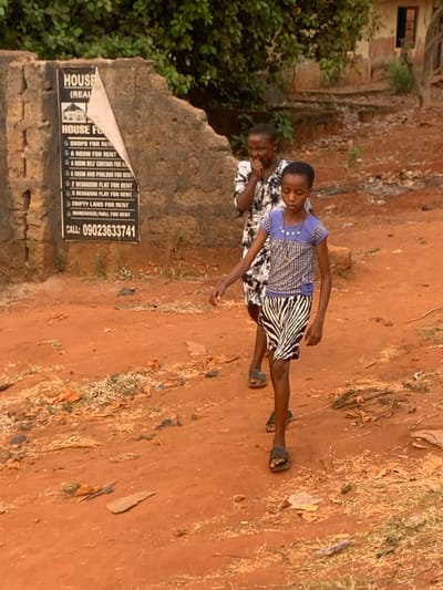 A woman and child stand on a dirt road near a weathered wall displaying a "House For Rent" sign. The sign lists various rental options, from shops to apartments, and includes a phone number. The woman behind the child looks toward the ground. The child, wearing a patterned dress, is in the foreground, facing the road. A building with greenery is visible behind the wall, and dry vegetation lines the edge of the road. The image appears to be taken in a rural setting.