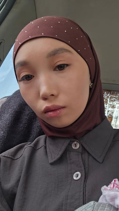 A close-up, selfie-style photograph shows a young woman wearing a brown hijab adorned with small, scattered rhinestones. She is also wearing a dark gray collared shirt with two visible buttons. The woman has fair skin, dark eyes, and is wearing makeup, including eyeliner and eyeshadow. Her lips are slightly parted, revealing a subtle pink lipstick. She is looking directly at the camera with a neutral expression. The background appears to be the interior of a vehicle, with a blurred gray seat visible to her left and a window with a bright, out-of-focus view to her left. The overall lighting suggests it is daytime. The woman is likely within the city of Kyzyl-Suu, Kyrgyzstan, given the context.