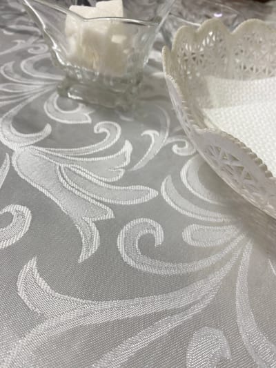 This is a close-up shot of a white damask tablecloth with a floral pattern. In the background, partially visible, is a clear glass dish containing several white sugar cubes, and a white decorative woven basket. The image appears to be an interior scene, likely a dining table setting. There is no discernible information about the city of Kyzyl-Kyya, Kyrgyzstan.