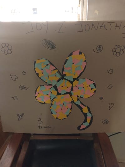 A handmade collage artwork on a light brown rectangular board is displayed vertically indoors in Barnawa, Nigeria. The central subject is a five-petaled flower with a curved stem, outlined in black. The flower's interior is filled with numerous small, irregularly shaped pieces of light blue, light pink, and yellow paper. Above the flower, the text "JOY . Z . JONATHAN" is written in black capital letters. Below the flower, the phrase "A Flower" is written in black script. Decorative elements, including two simple outlined flowers, multiple spirals, and several heart shapes, are drawn in black around the main flower. The artwork is positioned above a dark brown wooden chair with a black upholstered seat, visible in the lower frame. A blurred window displaying an outdoor scene with building structures provides background illumination above the artwork.