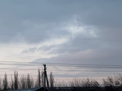 The image captures an overcast outdoor scene in what is identified as Argin, Uzbekistan. The upper two-thirds of the frame are dominated by a predominantly grey, cloudy sky with interspersed lighter, almost white, cloud formations. Multiple dark, horizontal utility lines span across the mid-ground of the image. A prominent, dark utility pole, appearing to be made of concrete or wood, is positioned in the mid-right, supporting several of these lines. Below the utility lines and the pole, a dense line of tall, bare deciduous trees forms a dark, silhouetted barrier against the horizon. In the lower-left corner, a dark, angled rooftop structure is partially visible. No individuals or discernable activities are present within the frame. The ambient light suggests either early morning or late afternoon conditions.