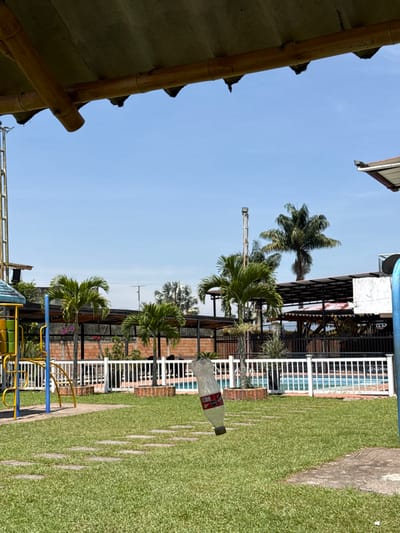 This image displays an outdoor recreational facility under a clear blue sky in Pitalito, Colombia. The upper portion of the frame features a bamboo and canvas canopy structure. The immediate foreground consists of a well-maintained green lawn intersected by a path of square stepping stones. A clear, partially crushed plastic bottle, identified as an empty soda bottle, is suspended in mid-air in the lower-right quadrant, oriented diagonally. To the left, a portion of a multi-colored children's playground structure, featuring yellow, blue, and green elements, is visible. A white picket fence extends horizontally across the midground, delineating the lawn from a swimming pool area where a blue pool is visible. Multiple small to medium-sized palm trees are interspersed throughout the midground, some planted within circular brick borders. In the background, buildings with brick walls and dark-roofed open-air sections are present, along with additional palm trees and a tall metal pole supporting a light fixture. No individuals are visible within the scene.