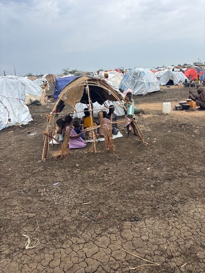 This image depicts a refugee camp in Al Babali, Sudan, under an overcast sky. The foreground is dominated by dry, cracked earth, with scattered dry grass and debris. In the center, a rudimentary shelter made of branches and woven material houses several individuals, including children. They appear to be seated or gathered within the shelter, possibly seeking respite from the elements or engaging in a communal activity.

Behind this central shelter, a densely packed area of tents and tarpaulins is visible, many of them appearing to be made of white, reflective material, some bearing the blue UNHCR (United Nations High Commissioner for Refugees) logo. The tents are arranged close together, indicating a crowded living situation. Scattered objects like plastic containers and buckets are also visible amongst the tents and on the ground. The scene suggests a humanitarian setting, likely a displacement camp. The overall impression is one of hardship and resilience. The time of day is difficult to determine precisely due to the overcast sky, but the lighting suggests it is daytime.