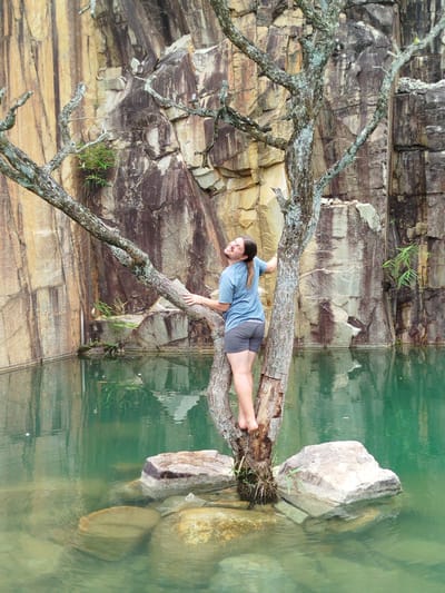 A person stands on a tree in a quarry in Lán Tranh, Vietnam. The person is wearing a blue t-shirt and grey shorts and is looking up with a smile. The tree is bare of leaves and is growing out of the water, which is a clear turquoise color. The quarry walls are made of rock and are a mix of brown and tan colors. There are several large rocks in the water around the tree. The scene is peaceful and serene.