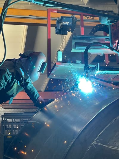 A welder, dressed in protective clothing and helmet, is welding a large, curved metal structure. The welding process is generating bright light and sparks. The welder is standing below the structure and reaching up to weld. A red and silver overhead crane-like structure with an electrical power unit is near the welding point. The environment appears to be an industrial setting. The image does not provide enough information to confirm the location in Zaljevo, Montenegro.