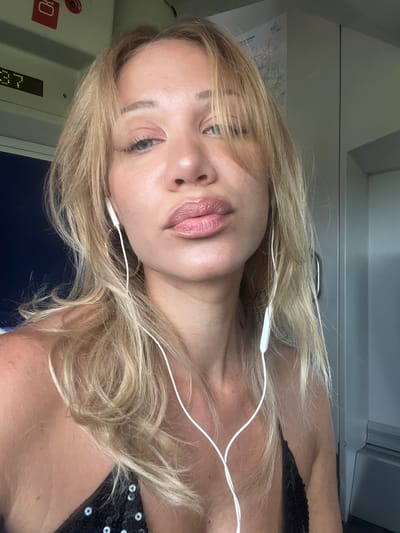 The media file is a close-up, slightly tilted selfie of a woman with blonde hair and fair skin. She is wearing white earbuds. Her lips are slightly parted and appear to have lip gloss on. Her eyes are open and looking towards the camera. Her hair is a light blonde color and appears to be slightly messy, falling around her face and shoulders. She is wearing a black top with visible sequins. The background appears to be the interior of a train, with gray walls and a dark blue seat visible. A map is affixed to the wall behind her. A red circular sticker with Cyrillic text, possibly "Ю", is visible on the left side of the frame, along with a digital display showing numbers that could indicate a time or route. The lighting suggests it might be daytime, but the interior setting makes it difficult to determine specific weather conditions. The overall mood is casual and relaxed. The image is a portrait of the woman, with the environment serving as a backdrop. There is no discernible activity taking place other than the woman taking a selfie.