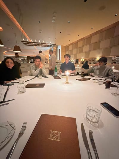 A restaurant table is set with white linens, glasses, cutlery, and menus. People are seated around the table, appearing to be in a dining setting. A lit candle provides soft illumination. Modern lighting fixtures are visible in the background. The overall ambiance suggests a formal or upscale restaurant.