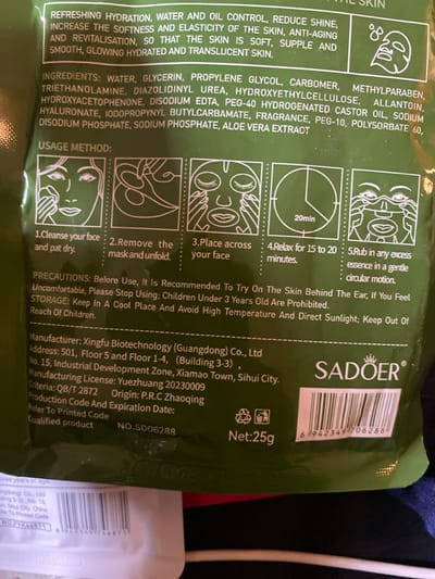 The image displays a close-up of a green, rectangular product package, likely for a face mask, oriented diagonally from the bottom left to the top right. The packaging is predominantly dark green with white text and line-art illustrations. The background is blurred, showing dark, indistinct shapes, possibly other items or fabric.

The front of the package is rich with information, divided into several sections:

1.  **Product Benefits/Claims (Top Left):** A block of text describes the product's intended effects: "REFRESHING HYDRATION, WATER AND OIL CONTROL, REDUCE SHINE, INCREASE THE SOFTNESS AND ELASTICITY OF THE SKIN, ANTI-AGING AND REVITALISATION, SO THAT THE SKIN IS SOFT, SUPPLE AND SMOOTH, GLOWING HYDRATED AND TRANSLUCENT SKIN." An icon resembling a face with water droplets is next to this section.

2.  **Ingredients List (Middle Left):** A detailed list of ingredients is provided: "WATER, GLYCERIN, PROPYLENE GLYCOL, CARBOMER, METHYLPARABEN, TRIETHANOLAMINE, DIAZOLIDINYL UREA, HYDROXYETHYLCELLULOSE, ALLANTOIN, HYDROXYACETOPHENONE, DISODIUM EDTA, PEG-40 HYDROGENATED CASTOR OIL, SODIUM HYALURONATE, IODOPROPYNYL BUTYLCARBAMATE, FRAGRANCE, PEG-10, POLYSORBATE 60, DISODIUM PHOSPHATE, SODIUM PHOSPHATE, ALOE VERA EXTRACT".

3.  **Usage Method (Center Right):** Titled "USAGE METHOD:", this section presents five illustrated steps for applying the mask. Each step has a corresponding white line-art drawing inside a circle:
    *   "1.Cleanse your face and pat dry." (Illustration shows hands patting a face.)
    *   "2.Remove the mask and unfold." (Illustration shows hands unfolding a mask.)
    *   "3.Place across your face" (Illustration shows a mask applied to a face.)
    *   "4.Relax for 15 to 20 minutes." (Illustration shows a clock with "20min" and a relaxed face with closed eyes.)
    *   "5.Rub in any excess essence in a gentle circular motion." (Illustration shows hands rubbing the face.)

4.  **Precautions and Storage (Bottom Middle):** This section outlines important warnings and storage instructions: "PRECAUTIONS: Before Use, It Is Recommended To Try On The Skin Behind The Ear, If You Feel Uncomfortable, Please Stop Using; Children Under 3 Years Old Are Prohibited. STORAGE: Keep In A Cool Place And Avoid High Temperature And Direct Sunlight; Keep Out Of Reach Of Children."

5.  **Manufacturer and Product Information (Bottom Right):**
    *   The brand name "SADOER" is prominently displayed in large white letters with a crown-like logo above it.
    *   A barcode is visible to the right of the brand name, with the number "6942349706288".
    *   "Net:25g" is indicated, accompanied by two recycling symbols.
    *   Detailed manufacturer information: "Manufacturer: Xingfu Biotechnology (Guangdong) Co., Ltd", "Address: 501, Floor 5 and Floor 1-4, (Building 3-3) No. 15, Industrial Development Zone, Xiamao Town, Sihui City."
    *   Further details include: "Manufacturing License: Yuezhuang 20230009", "Criteria: QB/T 2872", "Origin: P.R.C Zhaoqing", "Production Code And Expiration Date: Refer To Printed Code", "Qualified product", and "NO.SD06288" (a product number).

The overall impression is a commercially packaged cosmetic product with comprehensive instructions and information printed directly on the pouch.