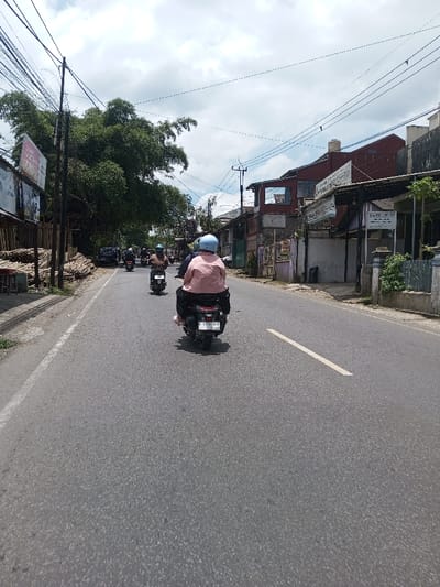 The image depicts a daytime street scene in Tagtse Tashidingkha, Bhutan, observed from a low angle, likely from a moving vehicle or scooter. The primary subject is a paved asphalt road with a solid white line on the left shoulder and a broken white line near the center, indicating lanes. Multiple individuals are riding scooters or motorcycles away from the camera. In the immediate foreground, a person wearing a light blue helmet and a pink shirt rides a dark scooter with a visible license plate: B 3318 UUY. Several other scooter riders, also wearing helmets, are visible further down the road.
On the left side of the road, a utility pole heavily laden with black wires stands next to a dense cluster of tall green bamboo foliage. Below the bamboo, a large billboard with partially visible text and images is mounted. Stacks of cut timber or logs are piled along the unpaved shoulder.
On the right side, a series of single and two-story buildings line the street. These structures vary in color and design, with elements suggesting both residential and commercial use. Several signs are affixed to the buildings, including one on a reddish building that reads "Praktek Dokter Umum." Utility poles with overhead wires also traverse this side of the road. The sky above is partially overcast with bright white clouds, indicating clear daytime conditions. No specific interactions between individuals are discernible, beyond the collective movement of traffic.