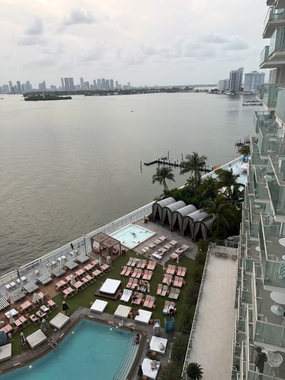 This is an elevated, wide-angle, over-the-shoulder shot of a resort pool area adjacent to a bay, with the Miami Beach skyline in the distance. The resort features multiple swimming pools, loungers, cabanas, and outdoor seating areas. The water is calm, and the sky is overcast. In the foreground, a portion of a modern apartment building with balconies is visible on the right. The Miami Beach skyline, featuring numerous skyscrapers, is visible across the bay on the left side of the image.